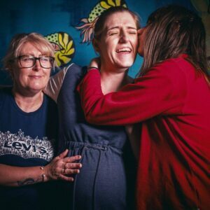 Three women embracing in front of a blue wall, showing love and friendship